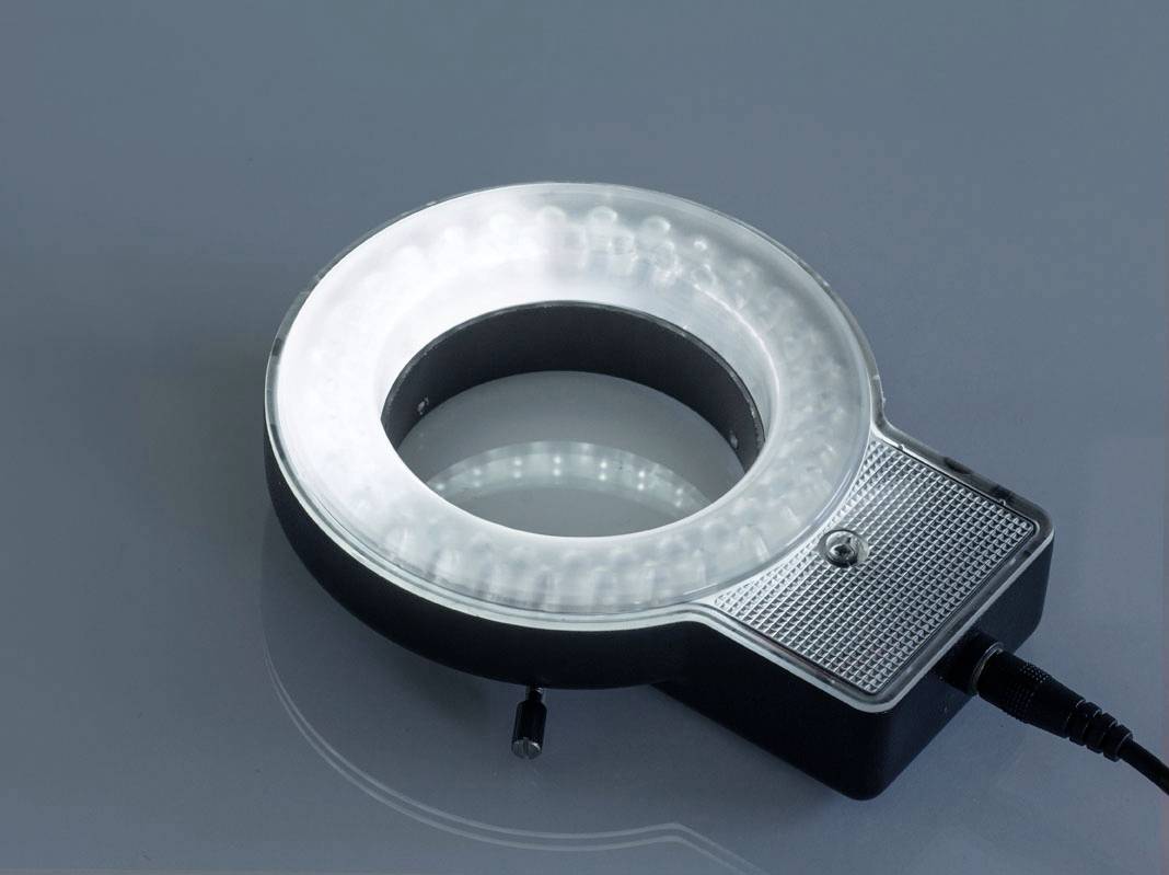 En rund LED-ring med plastskydd på grå bakgrund, ansluten via en kabel, sannolikt för fotograferingsbelysning.