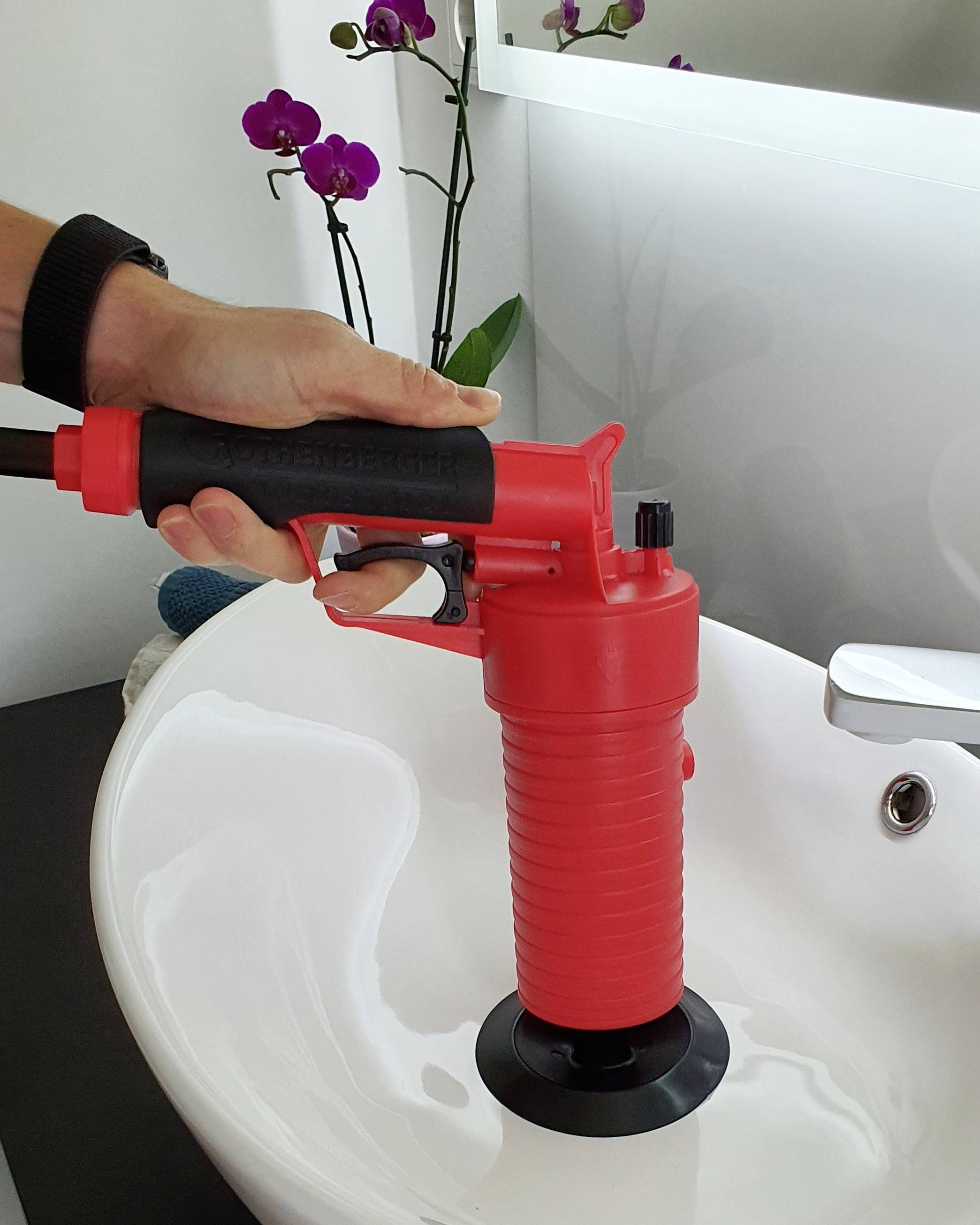 En hand använder en röd sugpropp för att rengöra ett vitt handfat. I bakgrunden står en orkidé på en hylla.