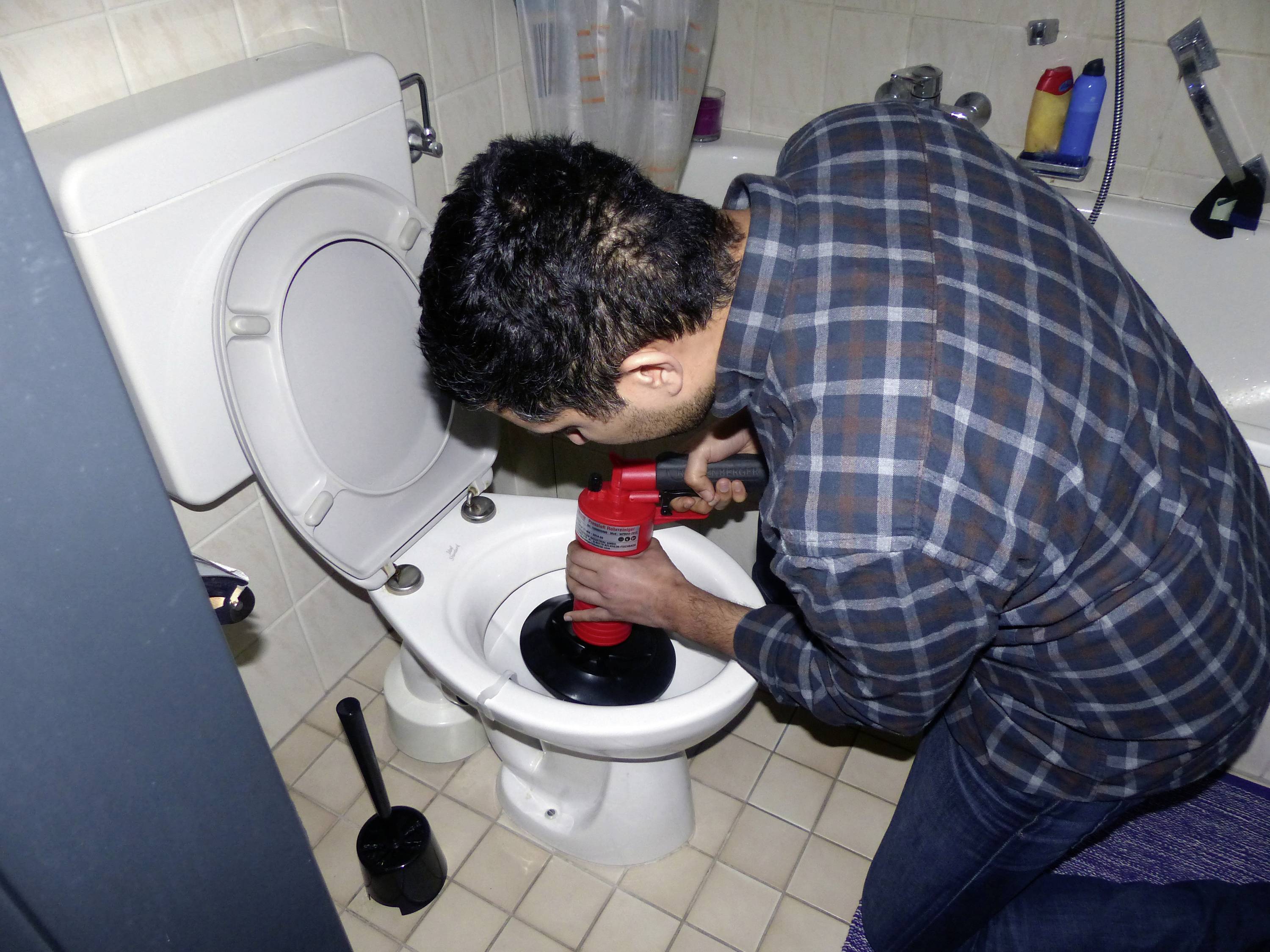 En person använder en sugpropp för att rengöra en igensatt toalett i badrummet. I bakgrunden syns rengöringsmedel.