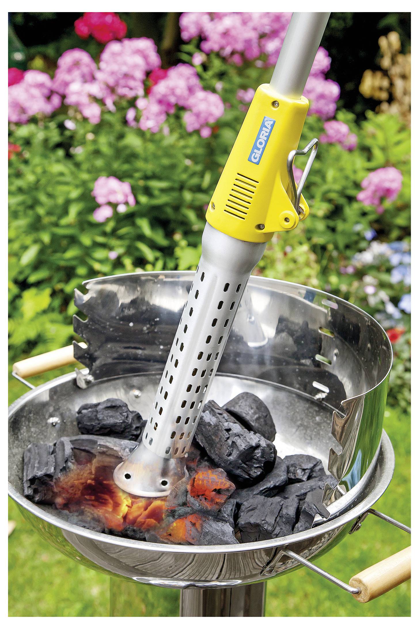Elektrisk kolstarter värmer kol i en rund utomhusgrill med glödande glöder och suddig trädgårdsblommor i bakgrunden.
