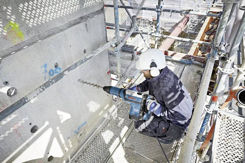 En arbetare i skyddsutrustning använder en elektrisk borr på en betongvägg på en byggarbetsplats, omgiven av byggnadsställningar, vilket belyser säkerhetspraxis.