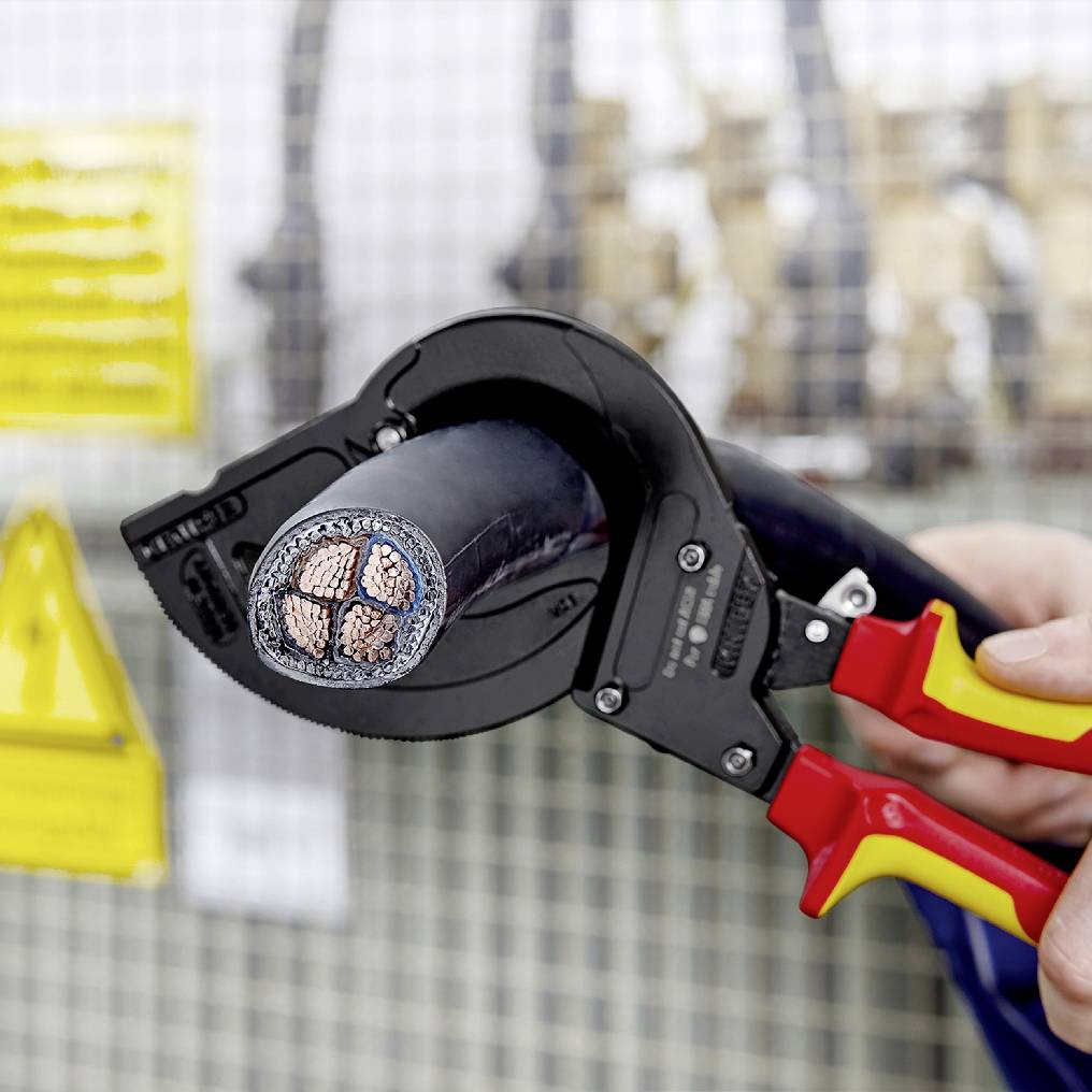 En kabelsax klipper av en tjock, bepansrad kabel. Skyltar i bakgrunden varnar för elektriska faror.