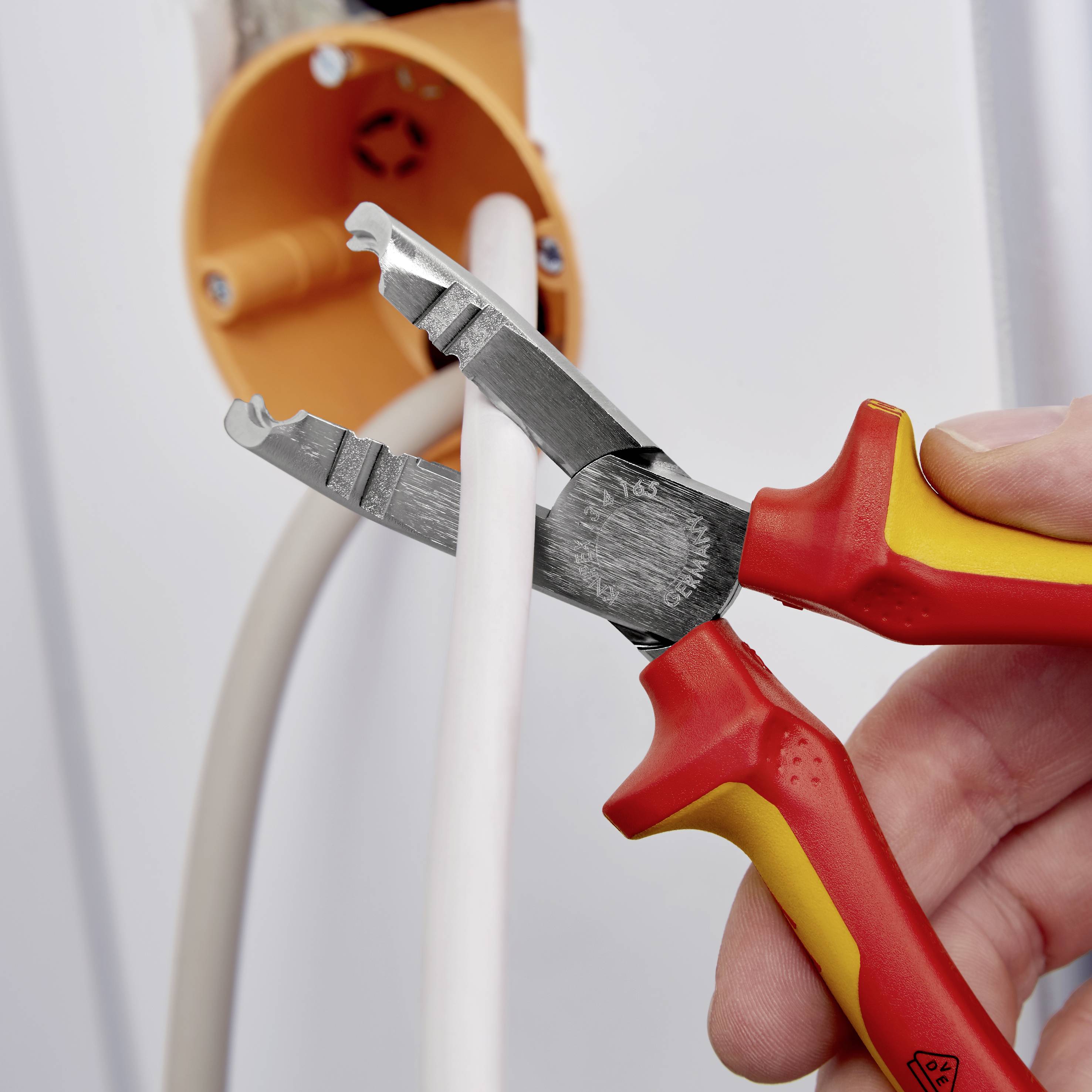 En hand håller en röd tång och klipper en elektrisk kabel i ett orange vägguttag.