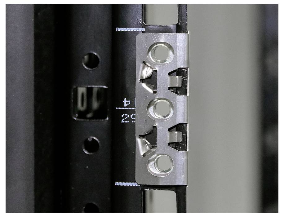 Närbild av ett metallband med stansade hål, fäst vid en svart yta. Tekniskt detalj i en industriell kontext.