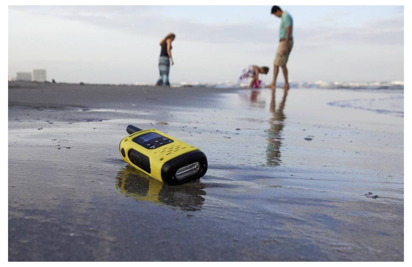 Gul handhållen walkie-talkie ligger i blöt sand på stranden, med sudiga människor i bakgrunden nära strandlinjen.