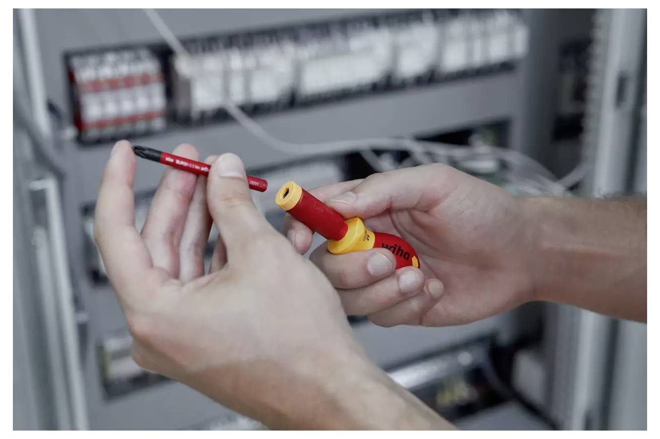 En person håller en röd och gul isolerad skruvmejsel framför en elskåp, vilket indikerar arbete eller underhåll på elektrisk ledning.