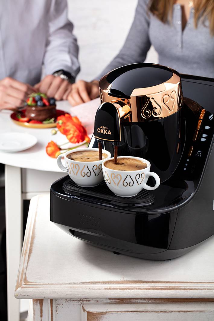 En snygg kaffebryggare tillagar två koppar kaffe samtidigt. I bakgrunden sitter två personer vid ett bord och äter.