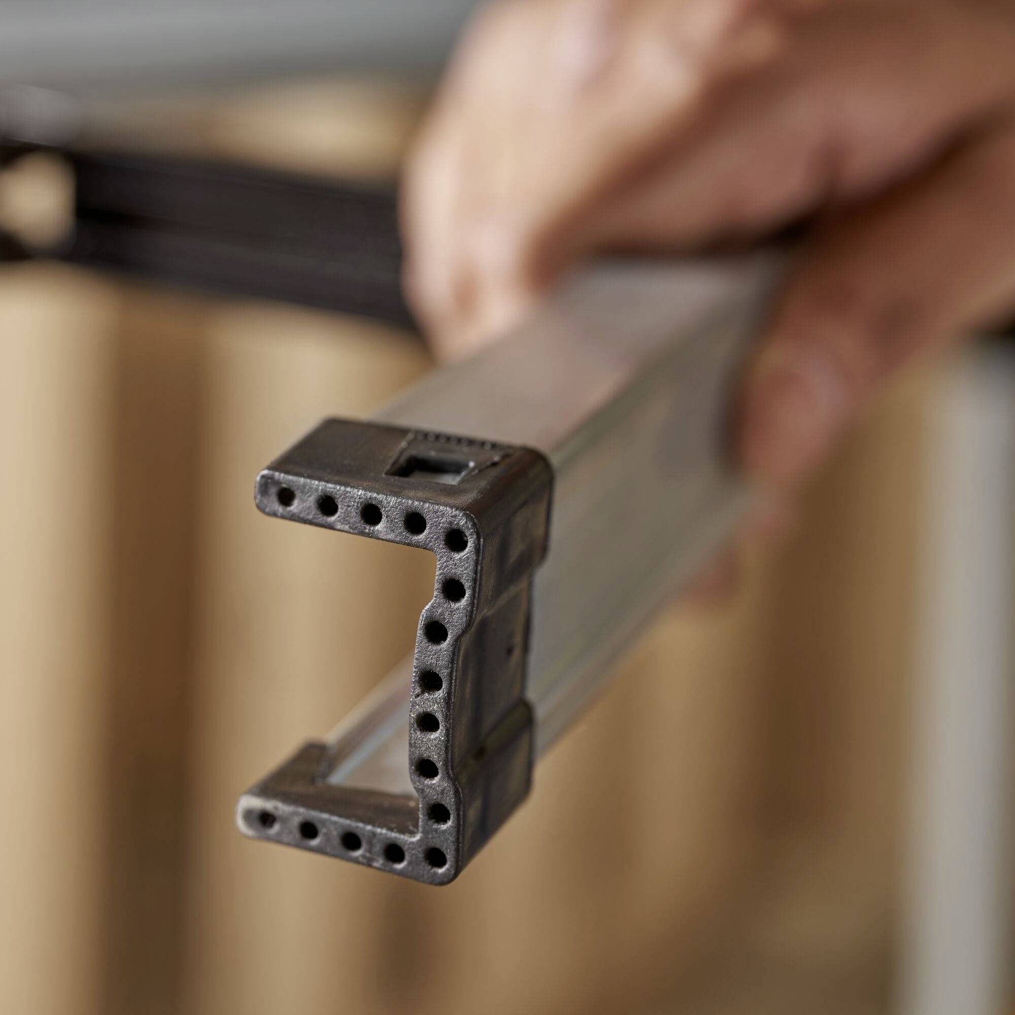 En hand håller ett metalliskt byggelement i profil, som verkar vara en del av en ram eller struktur.