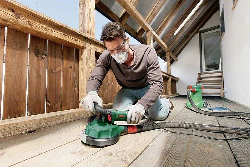 En man slipmar trätrallen på en balkongterrass på knä med en slipmaskin. Han bär handskar, skyddsglasögon och mask.