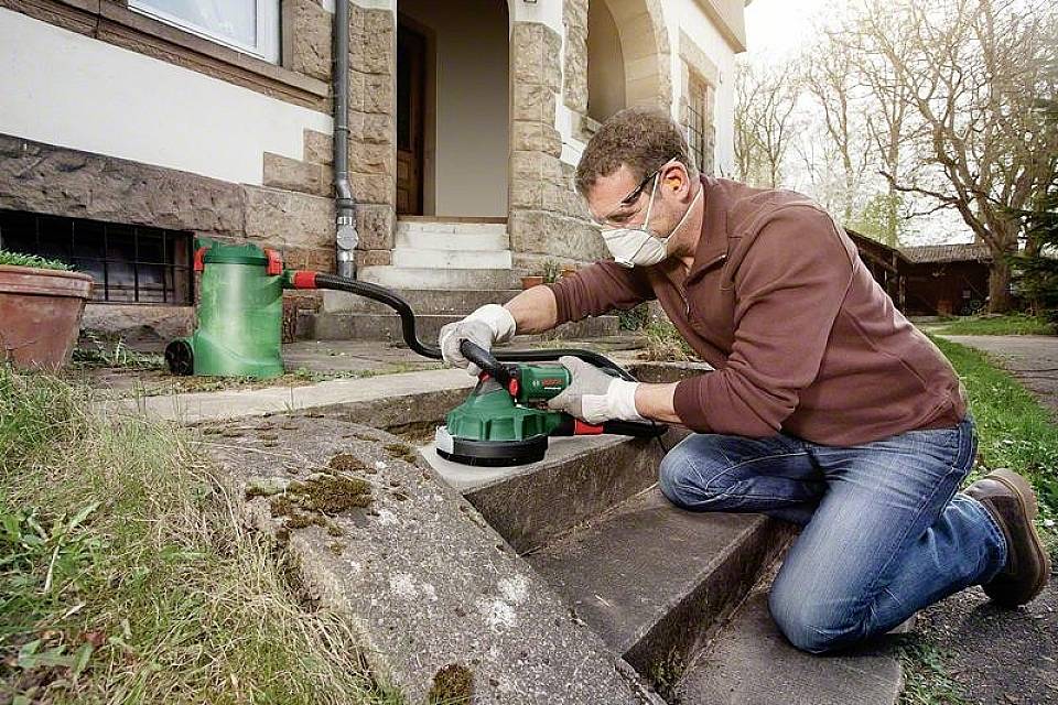 Man med skyddsglasögon och mask slipar stenytan på utomhustrappan. Dammsugare i bakgrunden i trädgården.