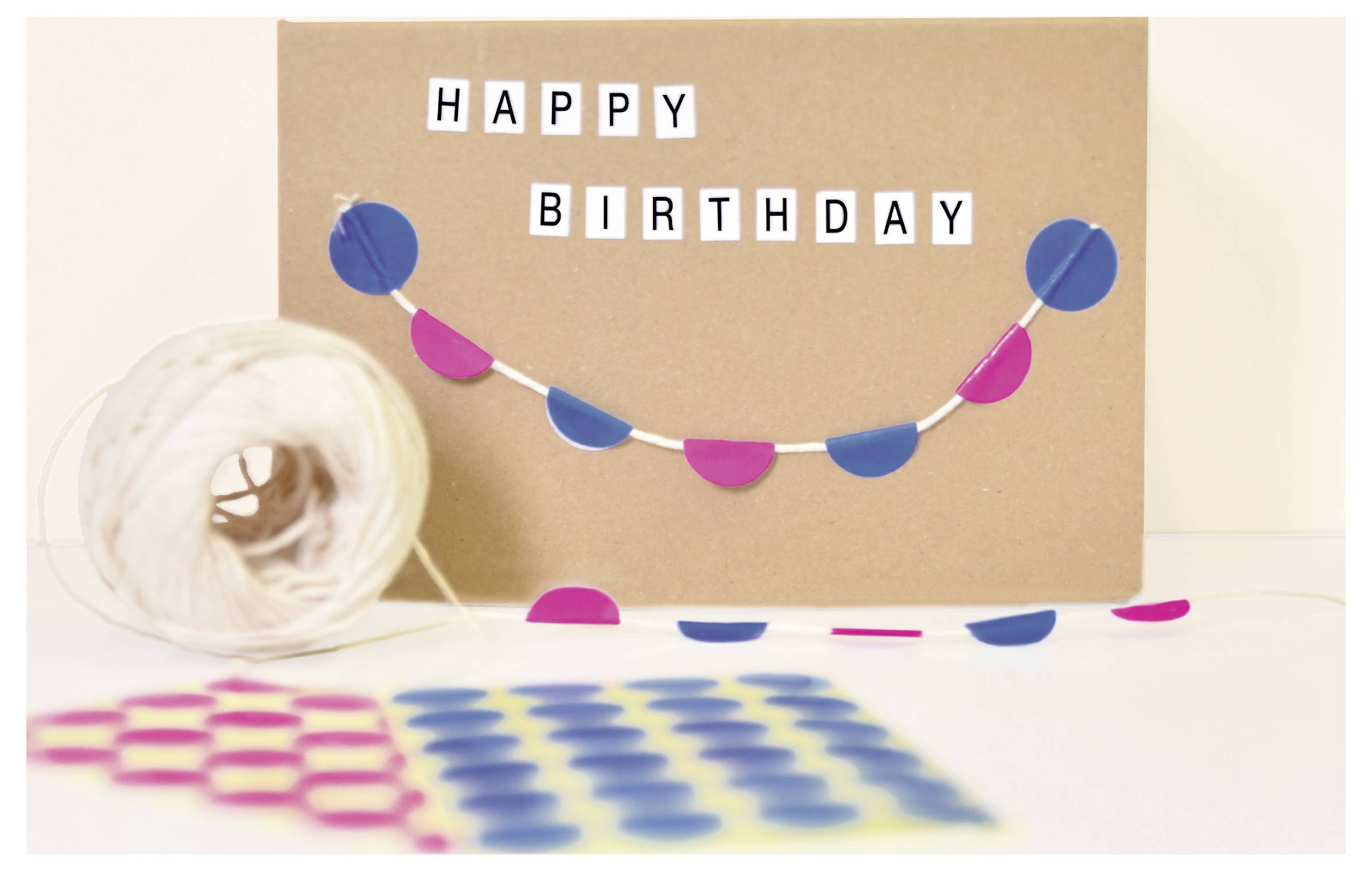 Födelsedagskort gjord av kraftpapper med texten 'HAPPY BIRTHDAY' och färgglad girlang; framför ligger pappersrullar och konfetti.
