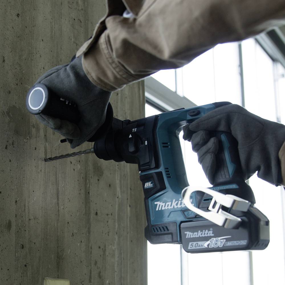 En person borrar ett hål i en betongvägg med en Makita borrhammare. I bakgrunden syns fönster.