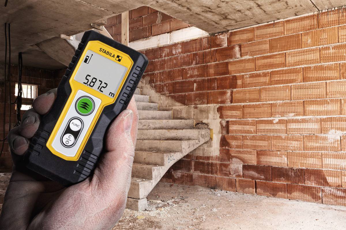 En hand håller ett avståndsmätningsinstrument som visar '5,872 m' framför ofärdiga trappsteg i en byggnad under konstruktion.