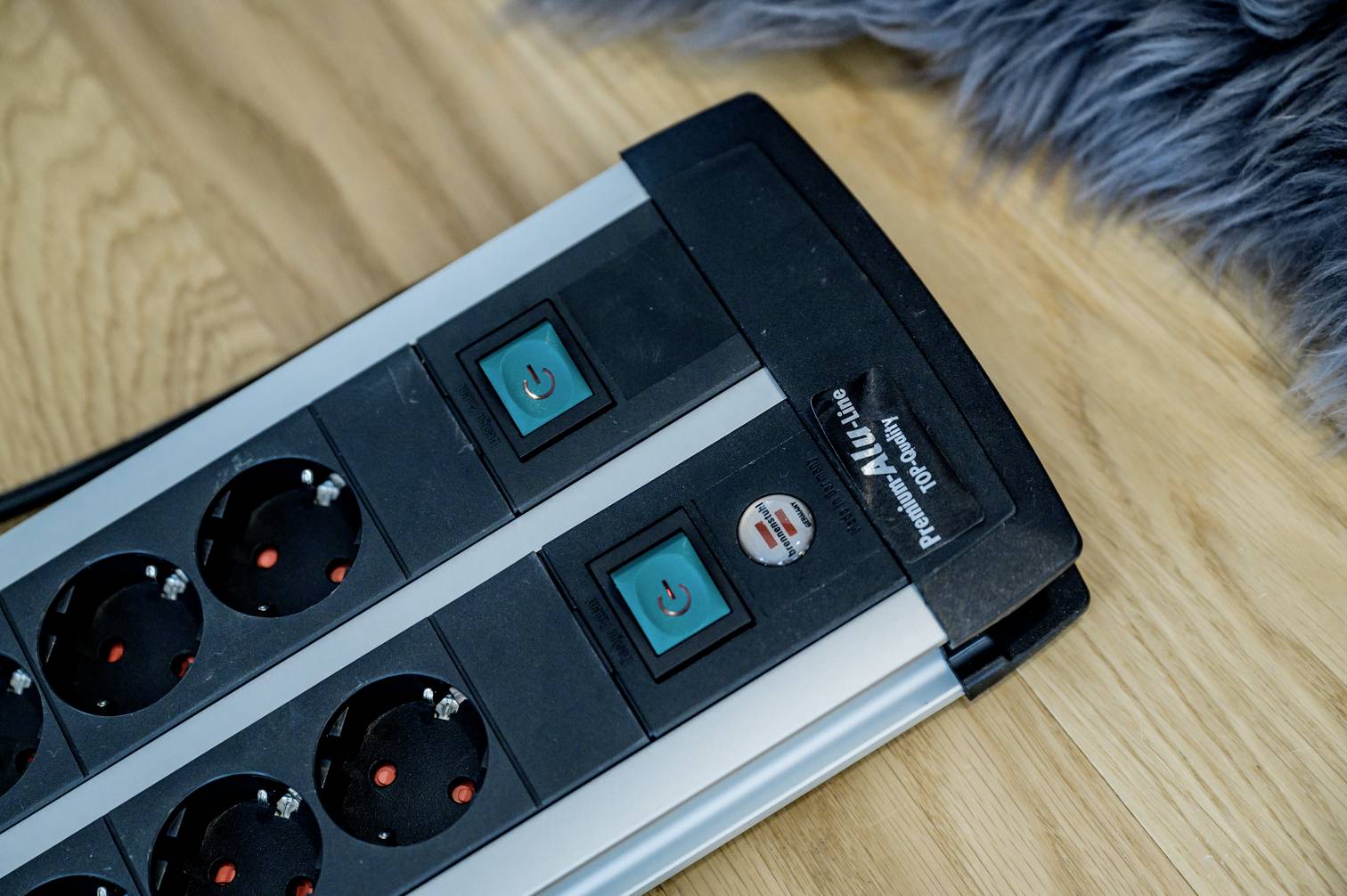 En grenuttag med strömbrytare ligger på ett trägolv. En del av en luddig grå matta syns i den övre bildkanten.