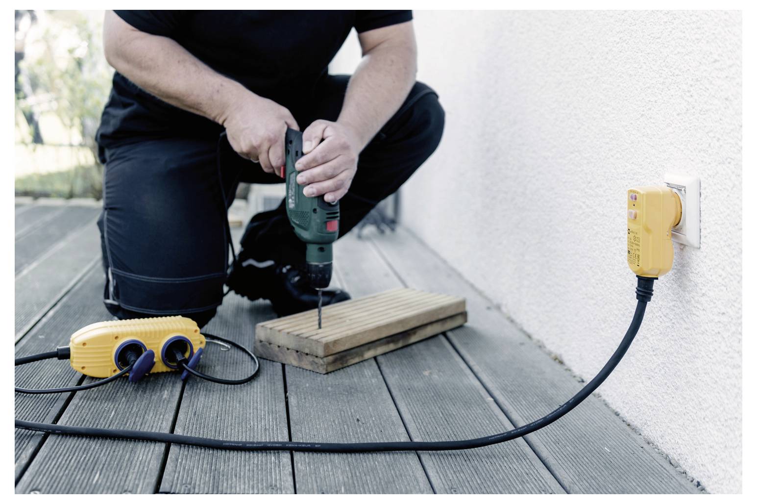 En person knäböjer på ett trädäck och borrar i en träbräda. En gul förlängningssladd och ett uttag är instickade i ett vägguttag i närheten.