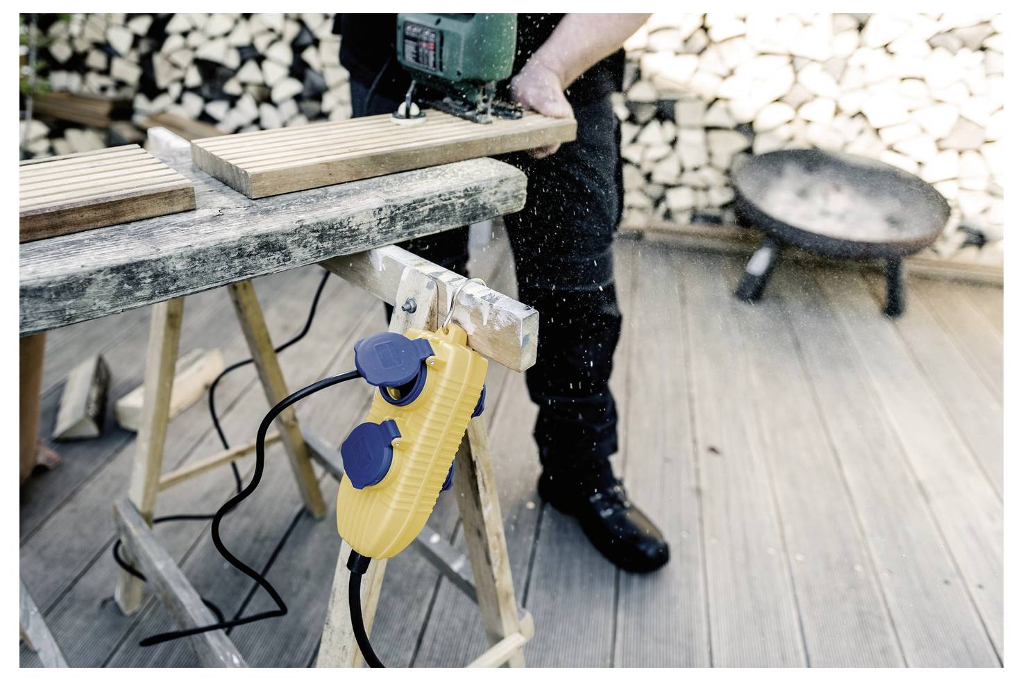 En person som använder en stickslip för att skära träplankor på ett såghäst utomhus, med staplade ved och en eldstad i bakgrunden.