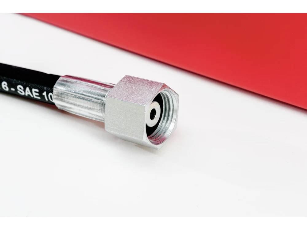 En metallisk hydraulisk slangkoppling som är fäst vid en svart slang ligger på en vit och röd yta, vilket indikerar industriell utrustningsanslutning.