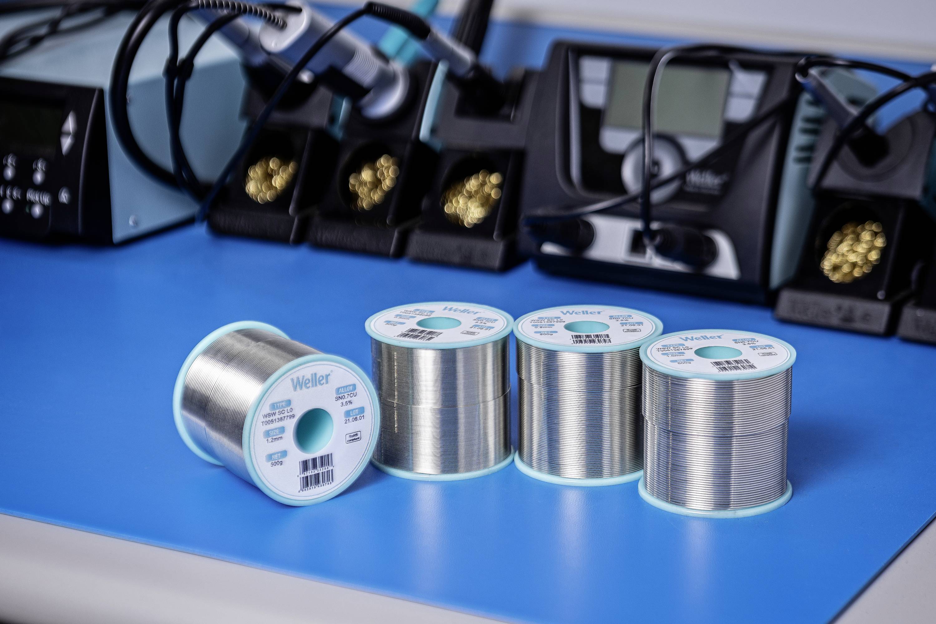 Fyra rullar lödtenn på ett blått bord, omgivna av lödstationer och lödkolvar. Perfekt för elektronik- och reparationsarbeten.