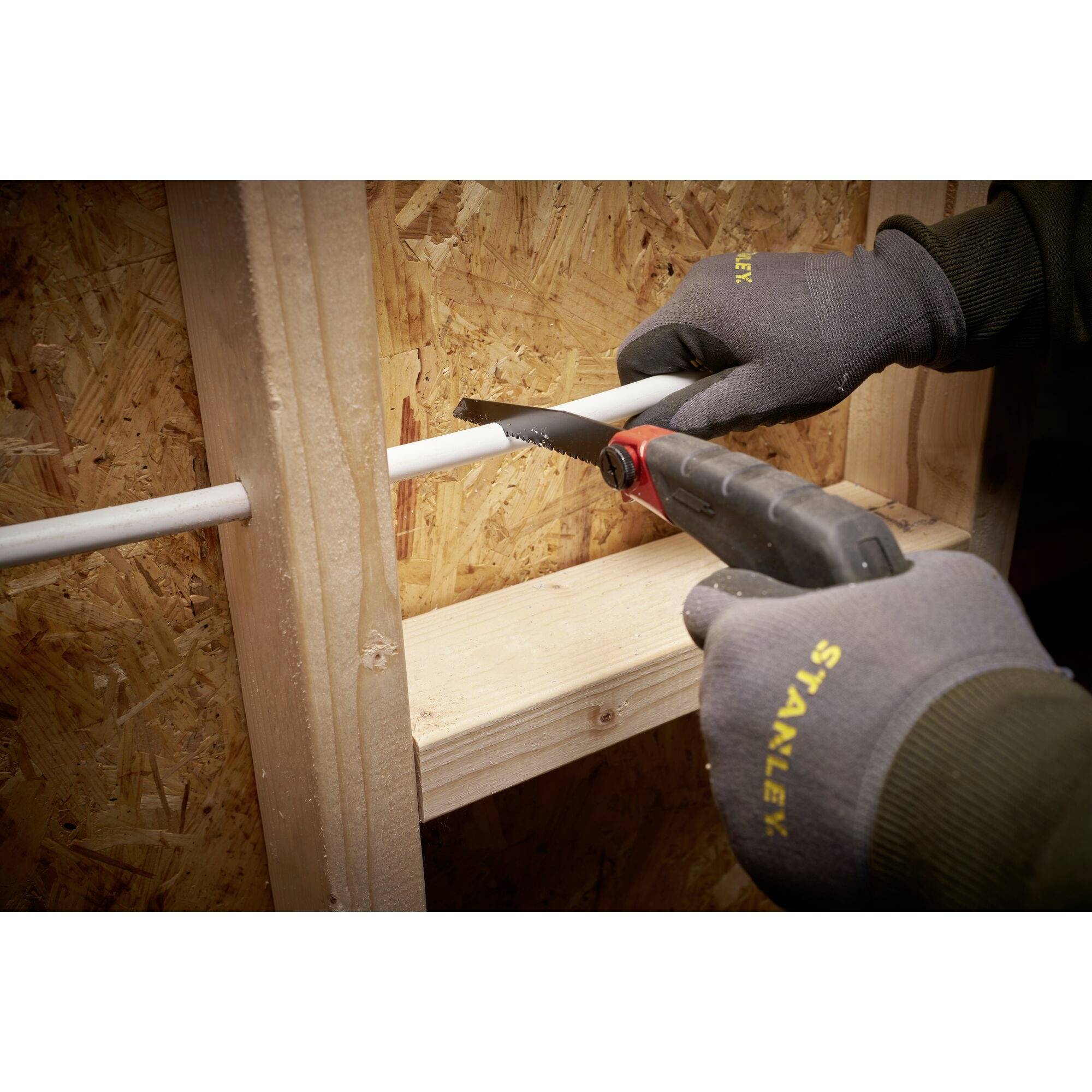 En person som sågar ett PVC-rör vid en trästruktur med en elektrisk såg bär skyddshandskar.