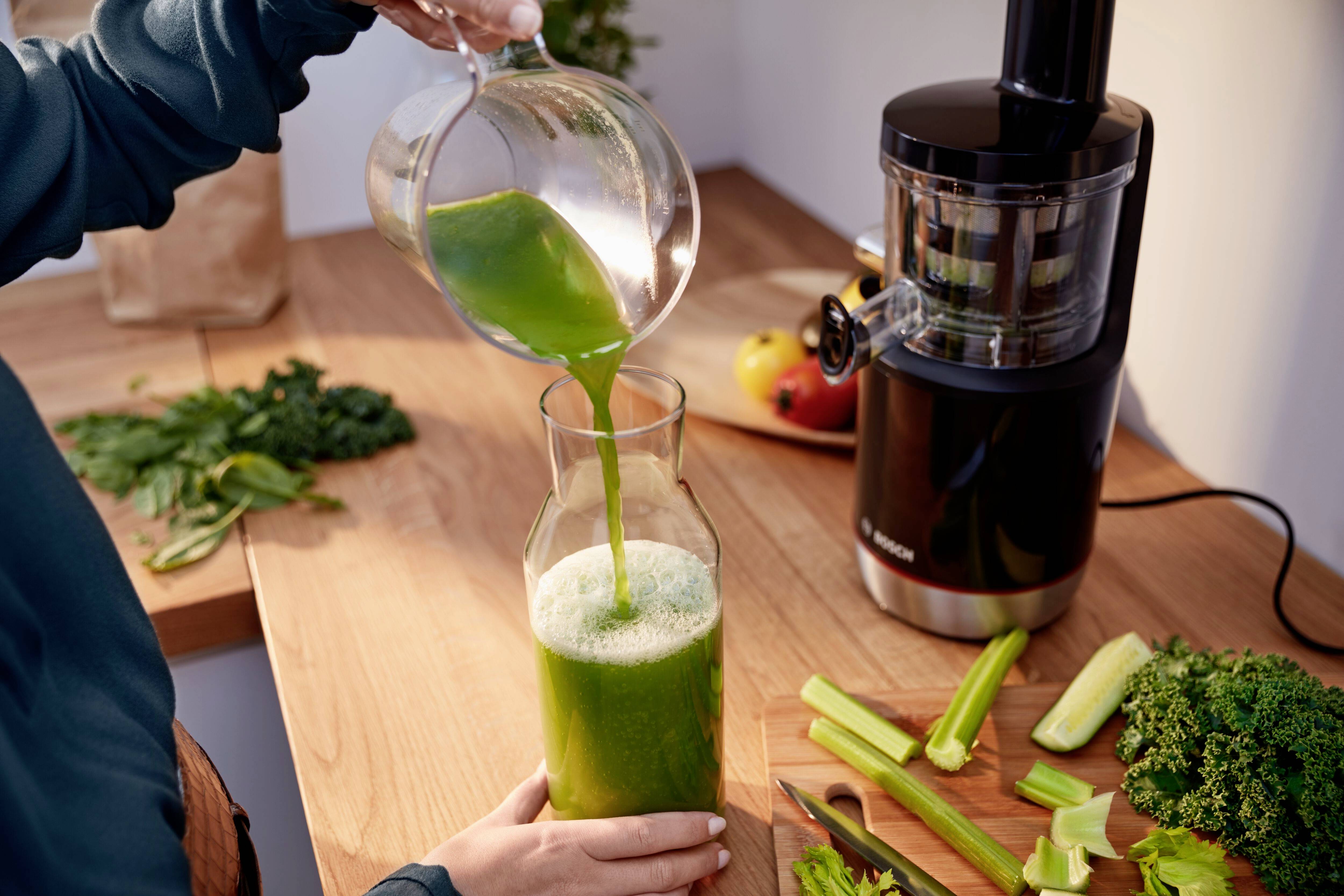 En person häller en grön smoothie i en glaskaraff på ett träarbetsbord. I bakgrunden syns frukt, grönsaker och en juicepress.