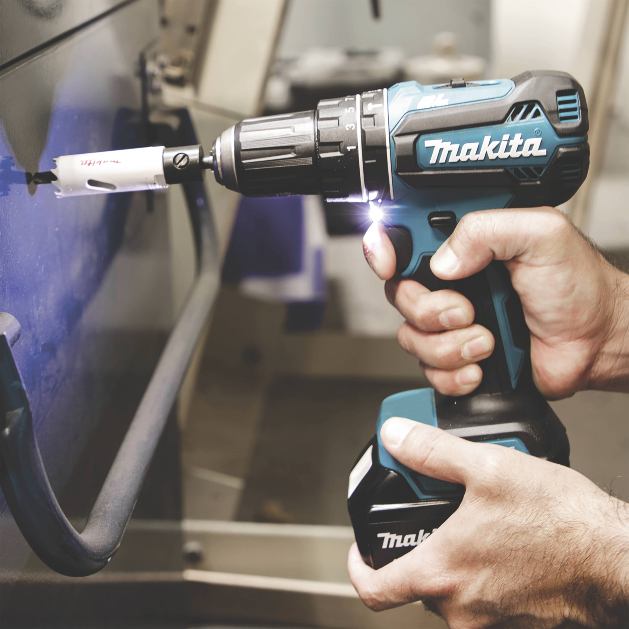 En person håller en blå Makita-borr med ett hålsågstillsats medan de bearbetar ett metallstycke.