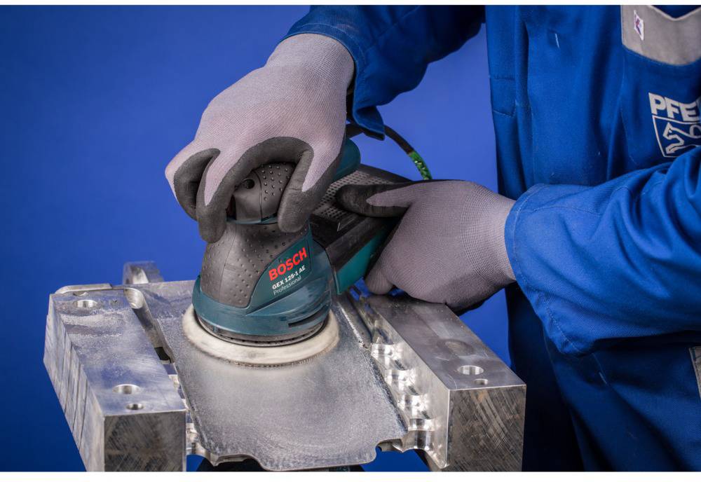 En person med arbetshandskar sliper en metalldetalj med en Bosch-slipmaskin mot en blå bakgrund.