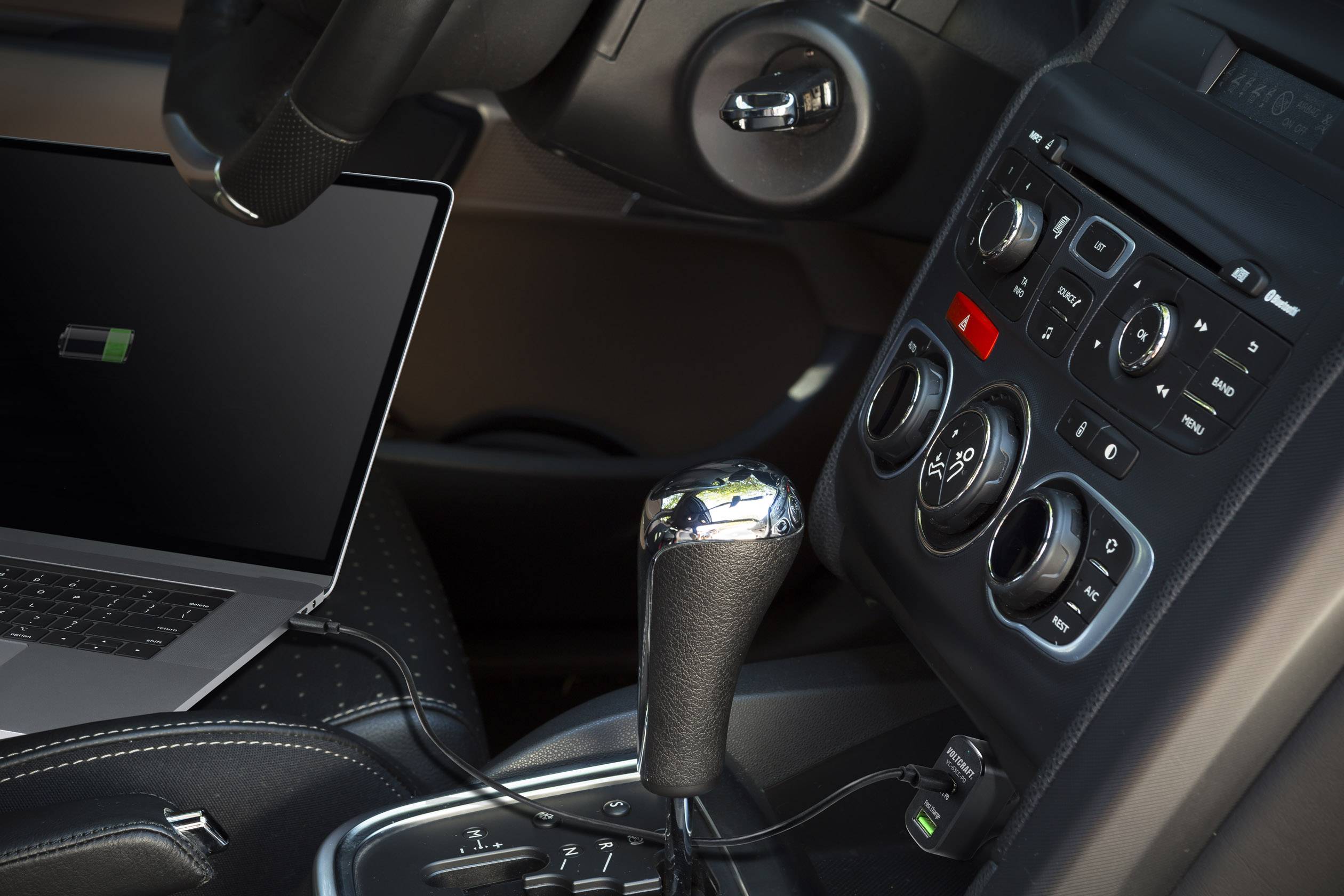 En laptop laddar i bilen, ansluten till instrumentbrädan. Synliga är ratten, mittkonsolen och växelspaken.