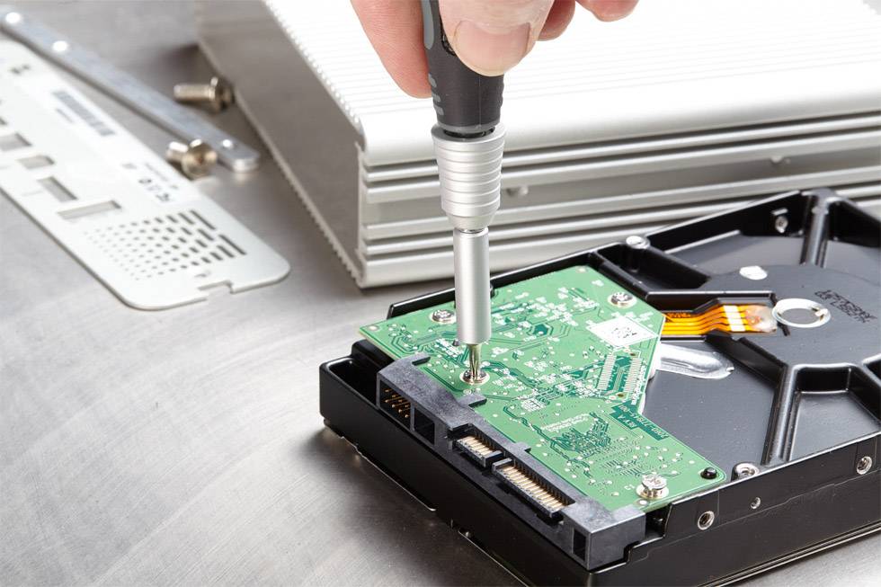 En person reparerar en hårddisk med en skruvmejsel. I bakgrunden finns ytterligare elektroniska komponenter.