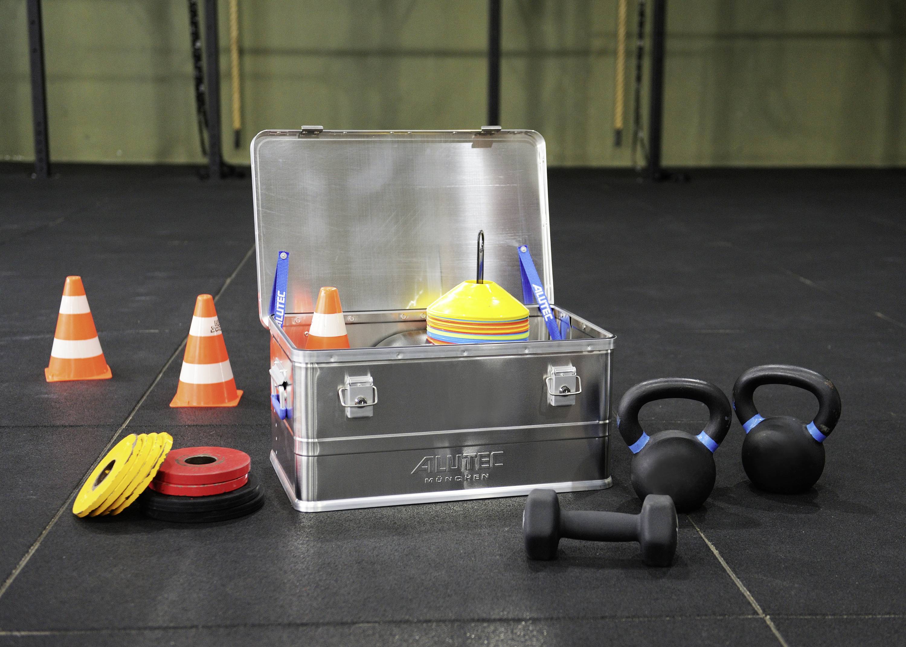 Träningsutrustning: Metallbox med hantlar, kettlebells, koner och viktplattor i en idrottshall, redo för ett träningspass.