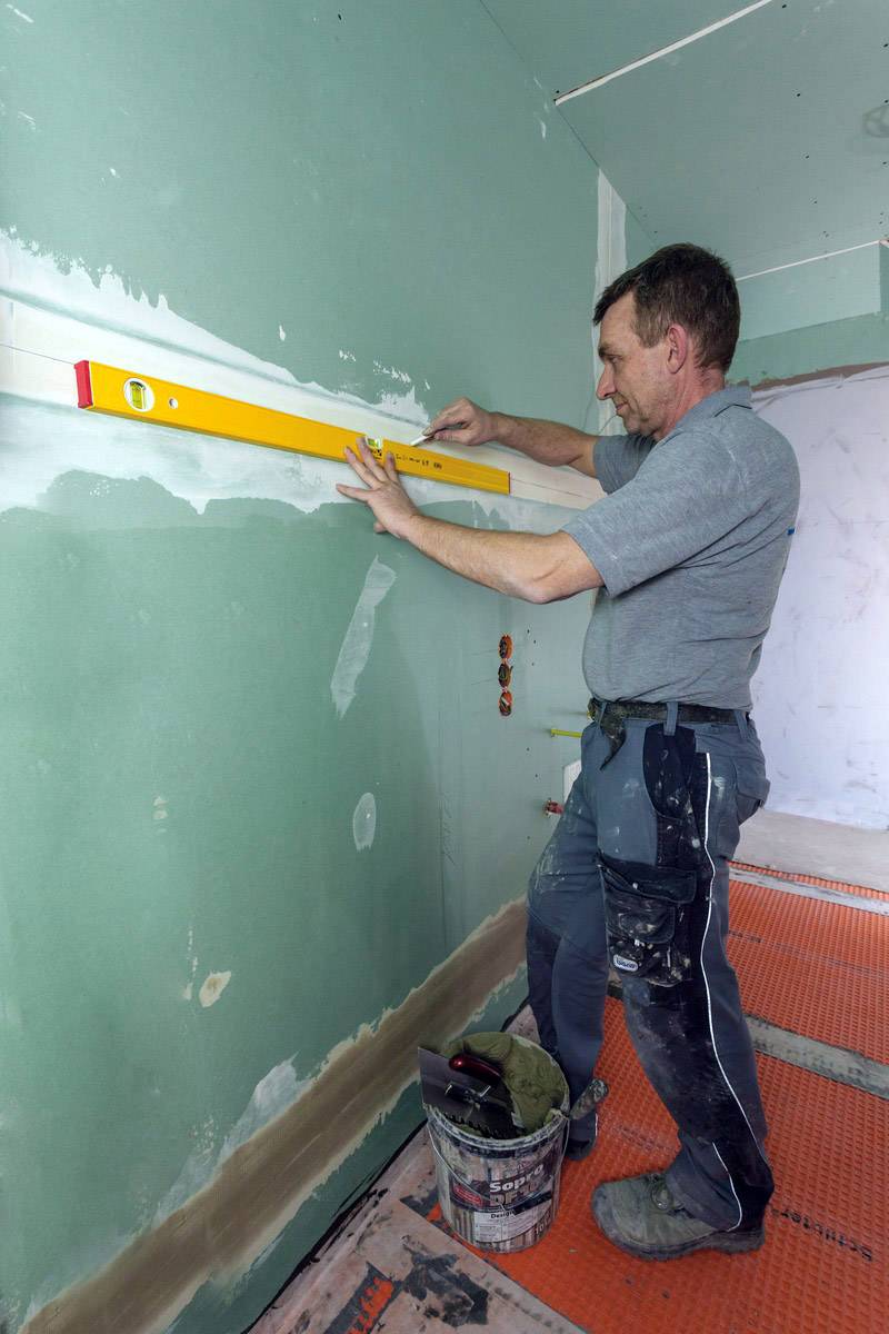 En hantverkare kontrollerar väggens planhet med ett vattenpass på en grön gipsskiva i ett rum som renoveras.