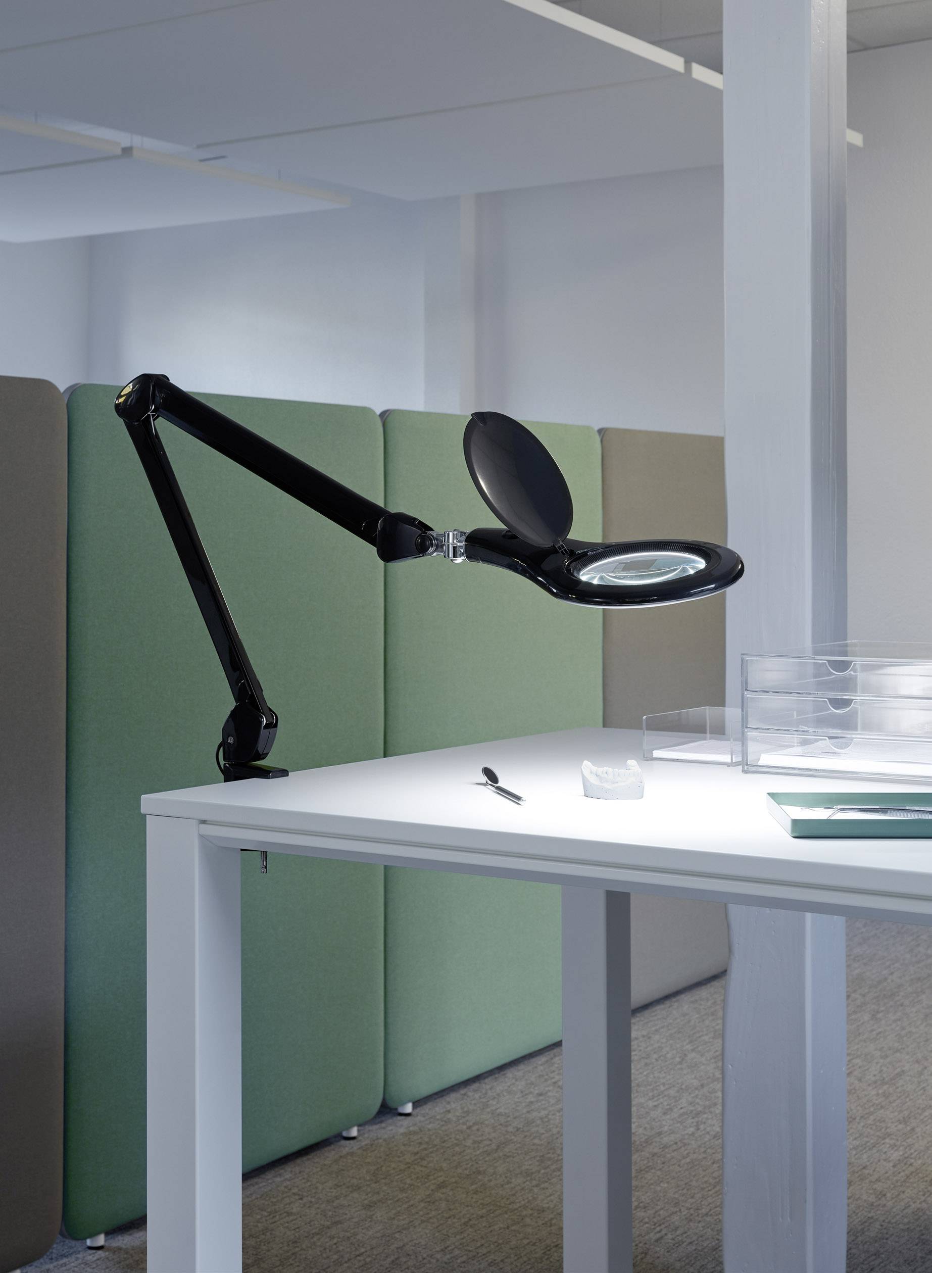 En lång, svart skrivbordsarm med förstoringsglas är placerad över ett ljust bord. I bakgrunden syns gröna skiljeväggar.