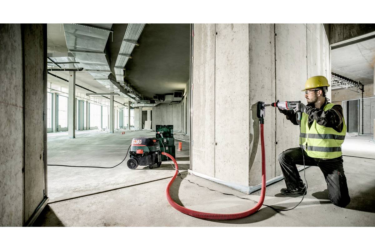 En byggnadsarbetare i skyddsutrustning använder ett elverktyg för att borra ett hål i en betongvägg. En dammextraktionsmaskin är ansluten.