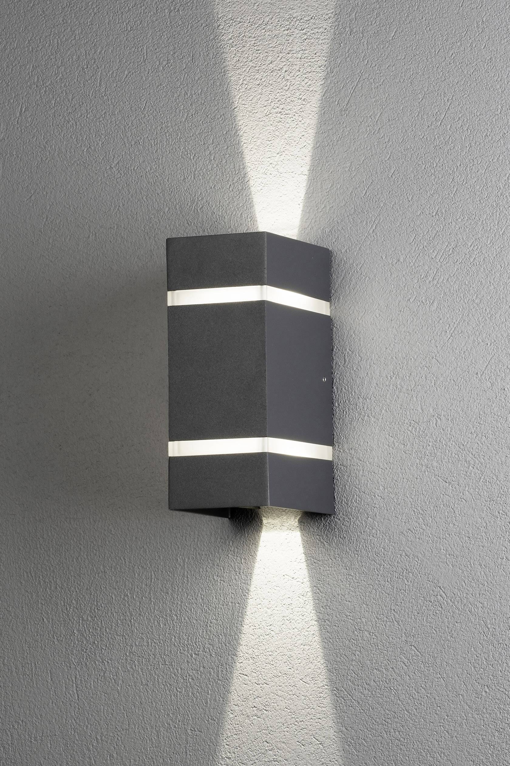 En modern, rektangulär vägglampa av mörkgrått metall lyser både uppåt och nedåt och skapar ett symmetriskt mönster på väggen.