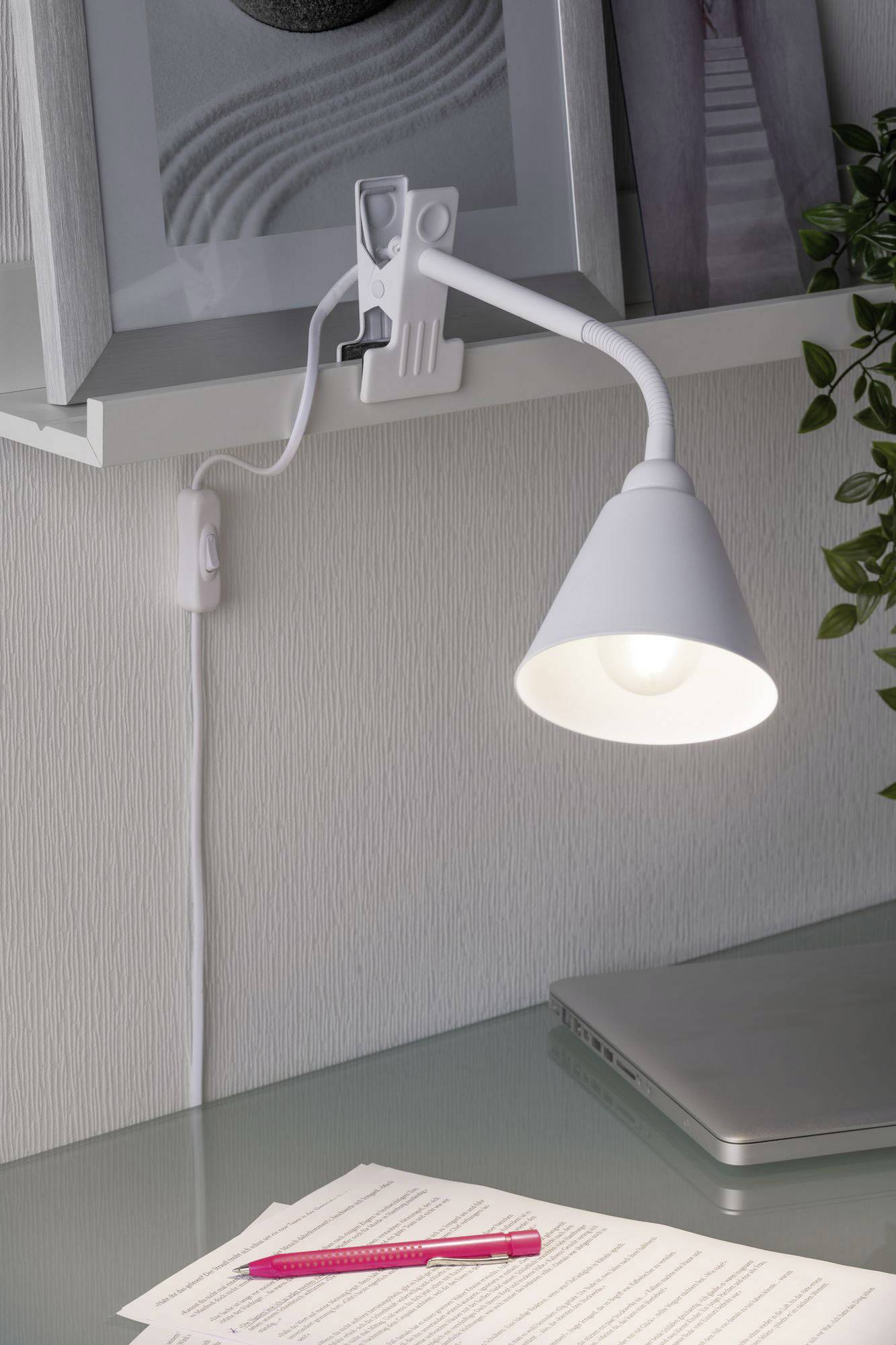 En vit klämlampa lyser upp ett skrivbord med dokument, en rosa penna och en bärbar dator. Lampan är fäst vid en hylla.