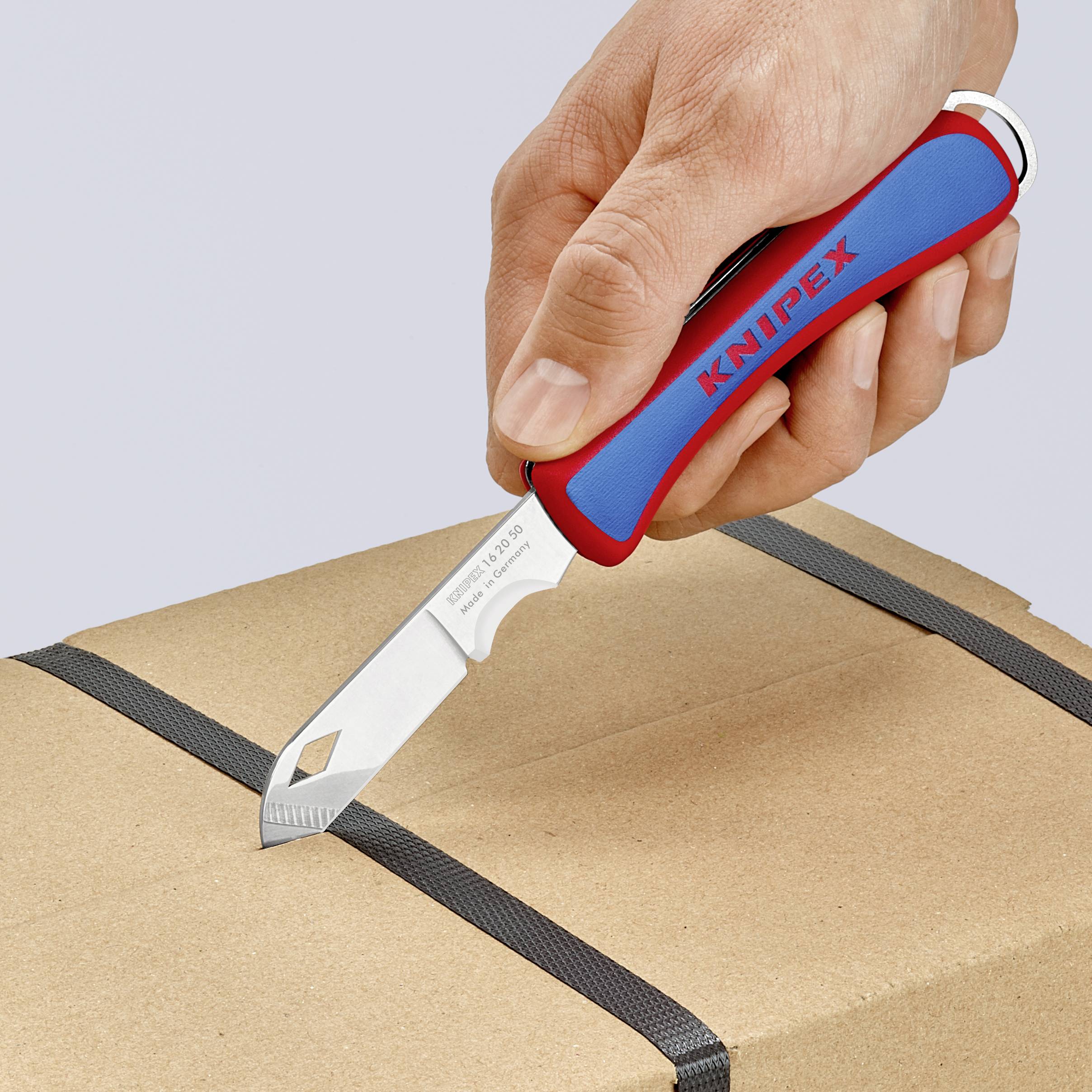 En hand håller en röd och blå skärkniv med texten 'Knipex' och klipper ett svart band på en kartong.