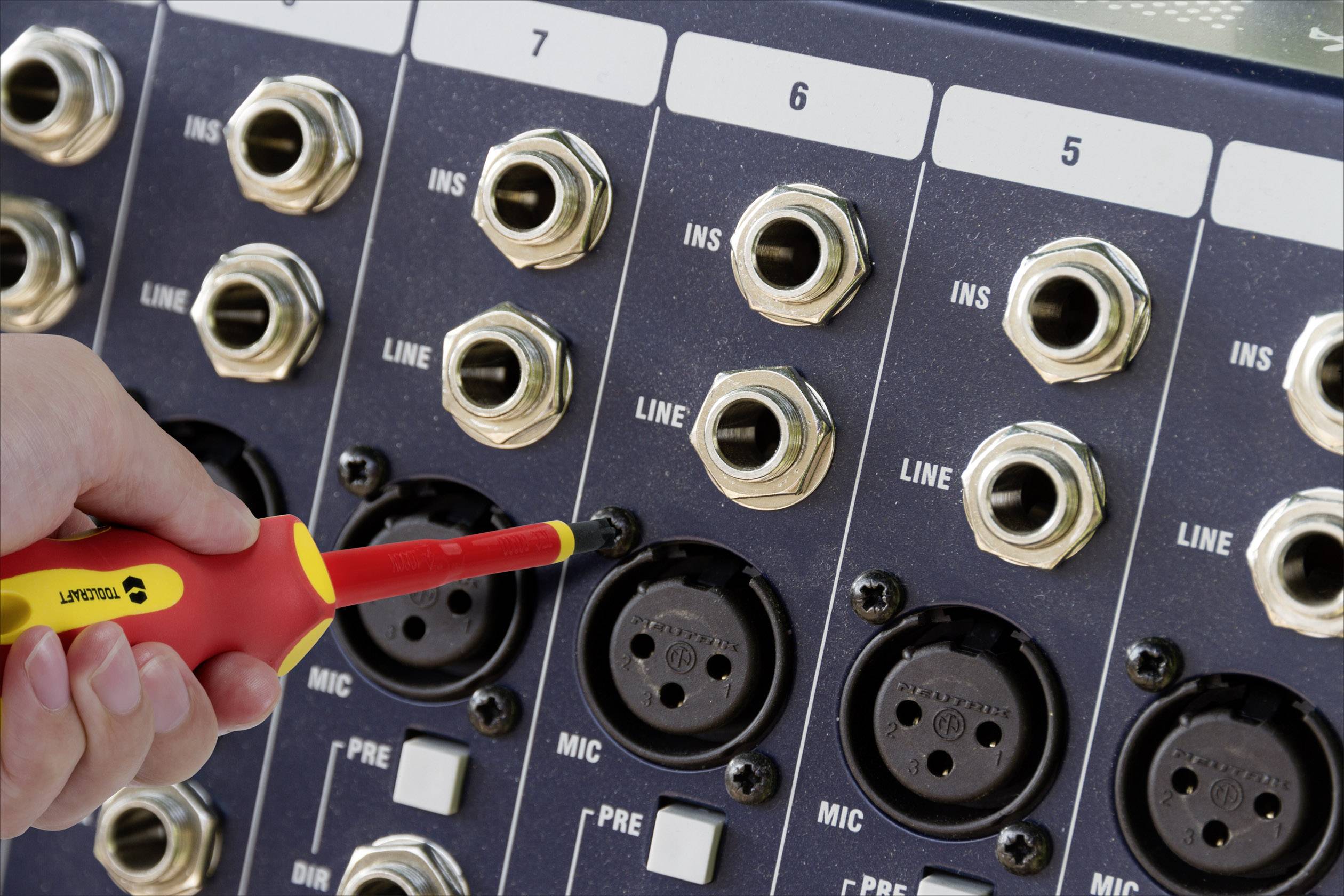 En hand sätter en röd skruvmejsel i en mikroingång på en audiopanel med flera kontakter och märkningar.