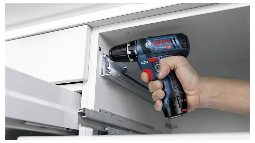 Hand som använder en sladdlös borrmaskin för att montera möbler, med fokus på att fästa metallskenor inuti ett skåp med precision.