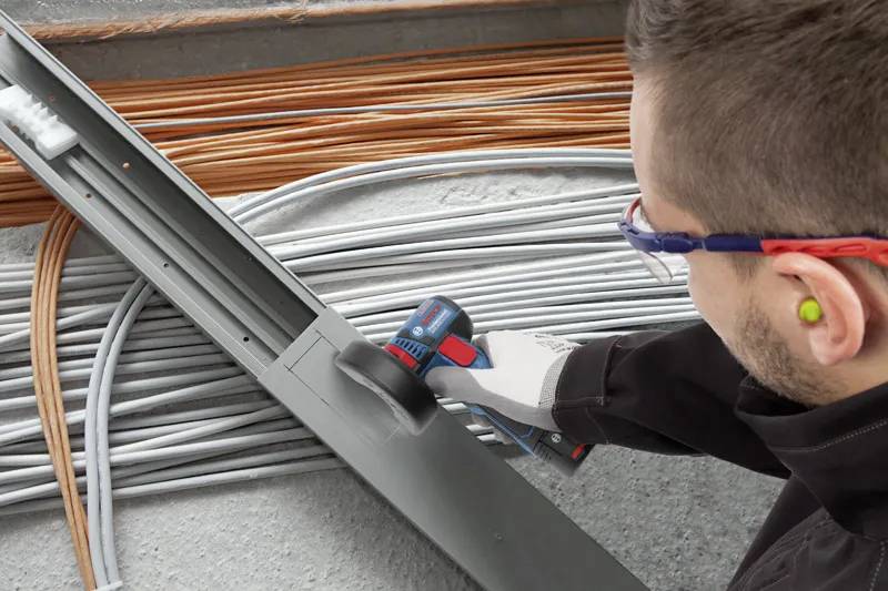 En person som använder ett elverktyg för att såga genom en metallbalk, omgiven av organiserade knippen av elkablar på en betongyta.