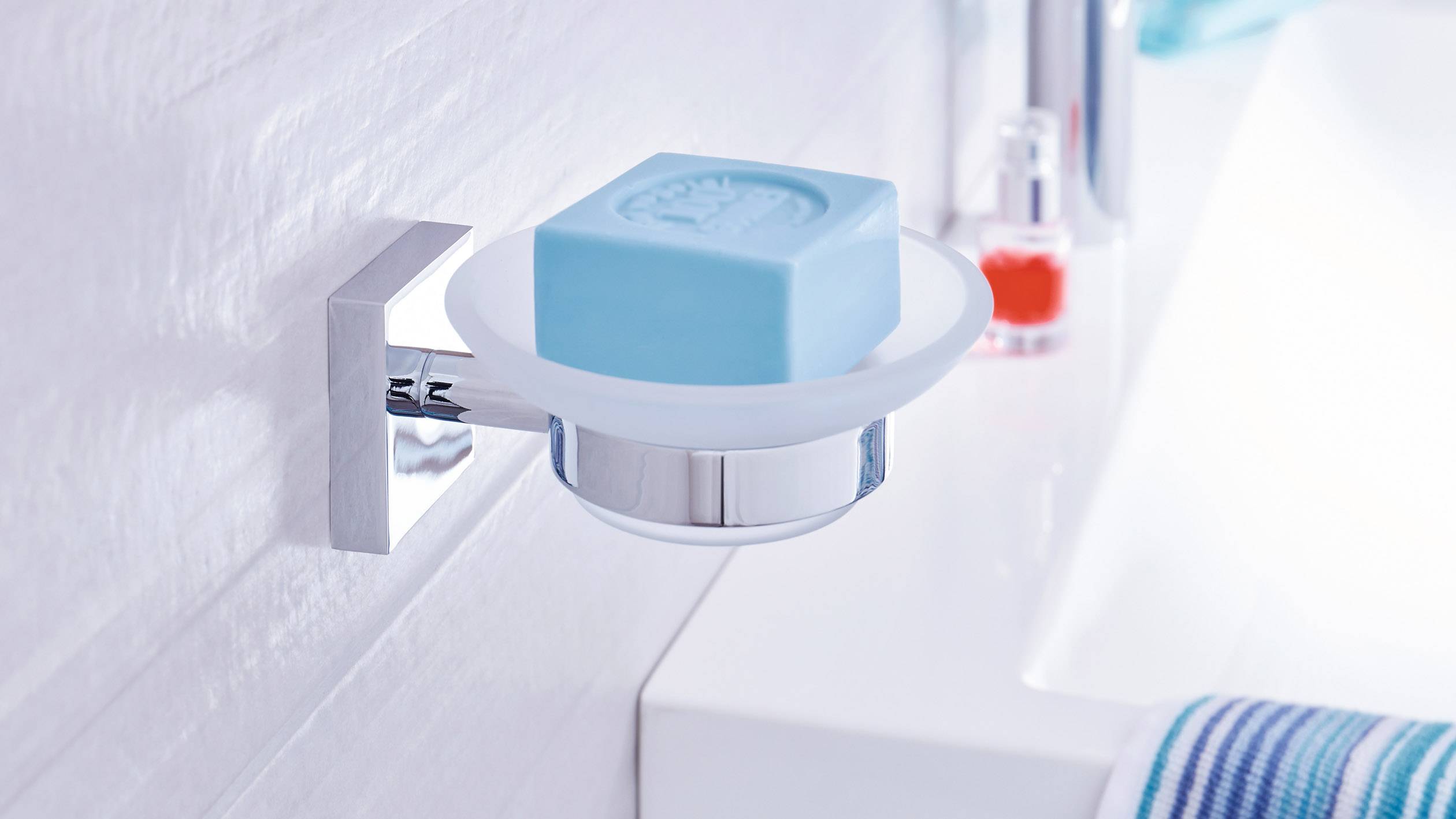 Blå tvål i en hållare på en badrumsmvägg, bredvid ett handfat med en handduk och en röd dispenser i bakgrunden.