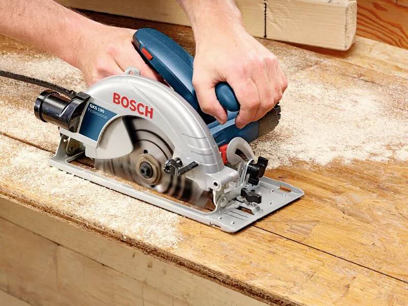 En person använder en Bosch cirkelsåg på en träarbetsbänk och sågar igenom ett trästycke med sågspån utspritt runt omkring.