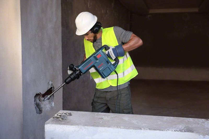 En byggnadsarbetare med skyddshjälm och varselväst borrar ett hål i en betongvägg med ett tungt elverktyg i ett oavslutat rum.