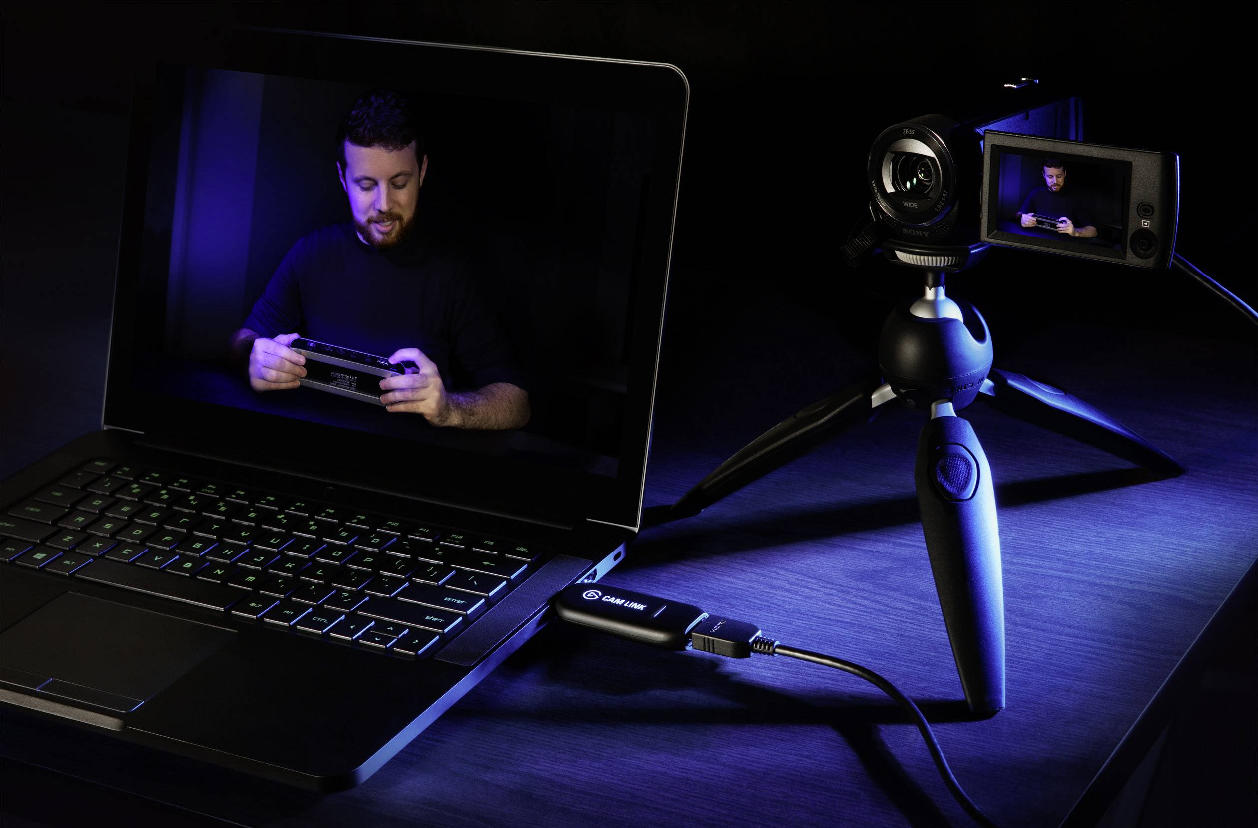 En laptop och en videokamera med stativ är anslutna. Laptopen visar en man som håller en enhet. Belysningen är mörk.