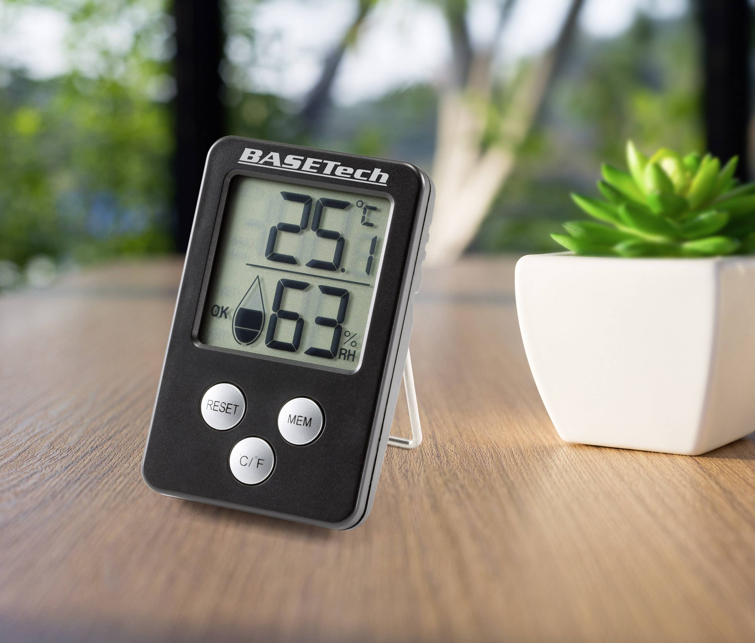 En digital termometer och hygrometer på ett bord visar 25,1 grader Celsius och 63% luftfuktighet. Bredvid står en liten växt.