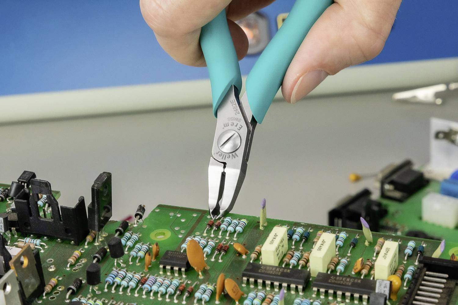 En hand håller en avbitare och klipper en tråd på ett grönt kretskort som är utrustat med olika elektroniska komponenter.