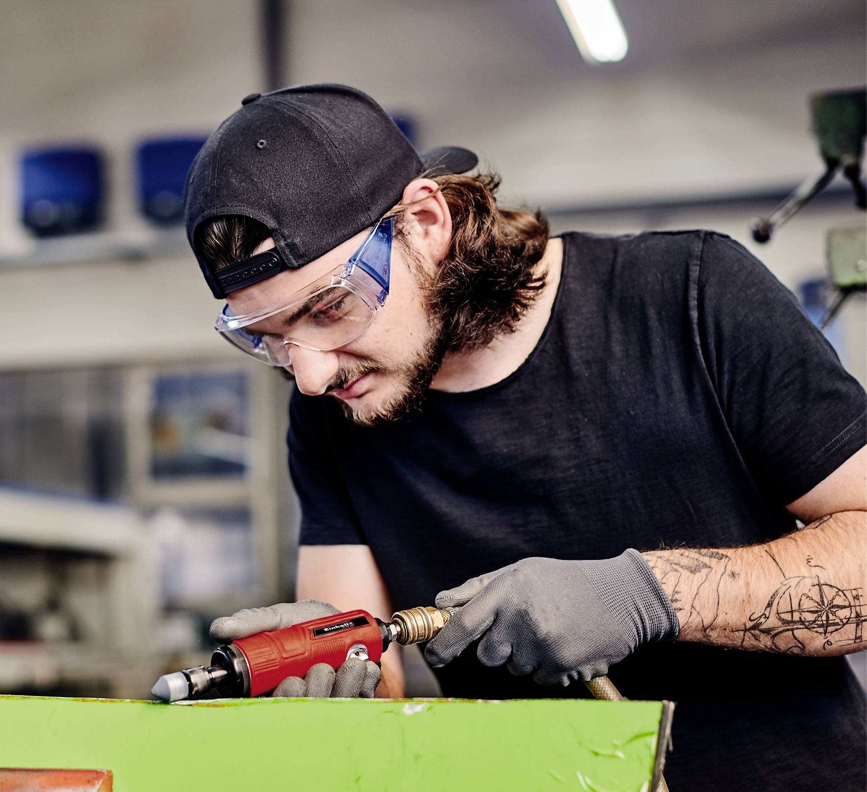 En person med skyddsglasögon och handskar använder ett elektriskt verktyg i en verkstad. Arbetsmiljön syns i bakgrunden.