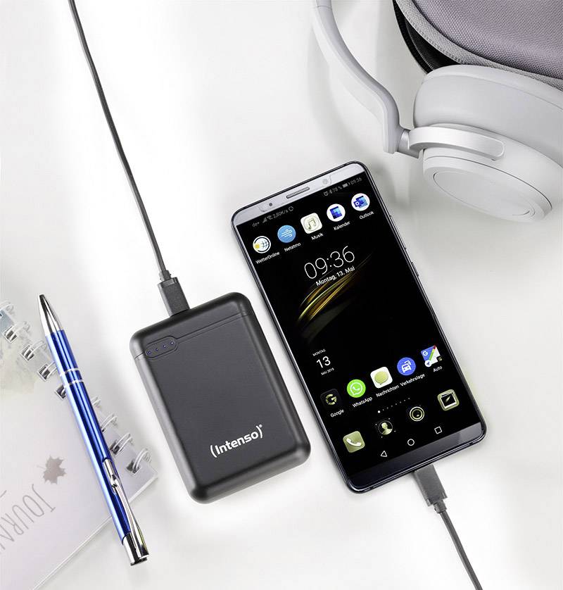 En smartphone och en "Intenso"-powerbank är anslutna med kablar och laddar. Bredvid ligger ett par hörlurar och en penna på ett bord.