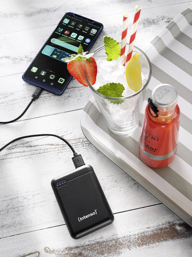 En smartphone laddas via en powerbank. Bredvid finns ett bord med ett glas fyllt med en iskallt dryck, jordgubbsskivor och en citronskiva, samt en juiceflaska.