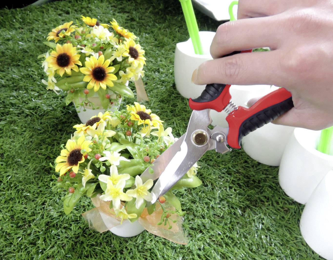 En hand klipper blommor med en sax i en liten, blommande kruka på en grön gräsmatta.
