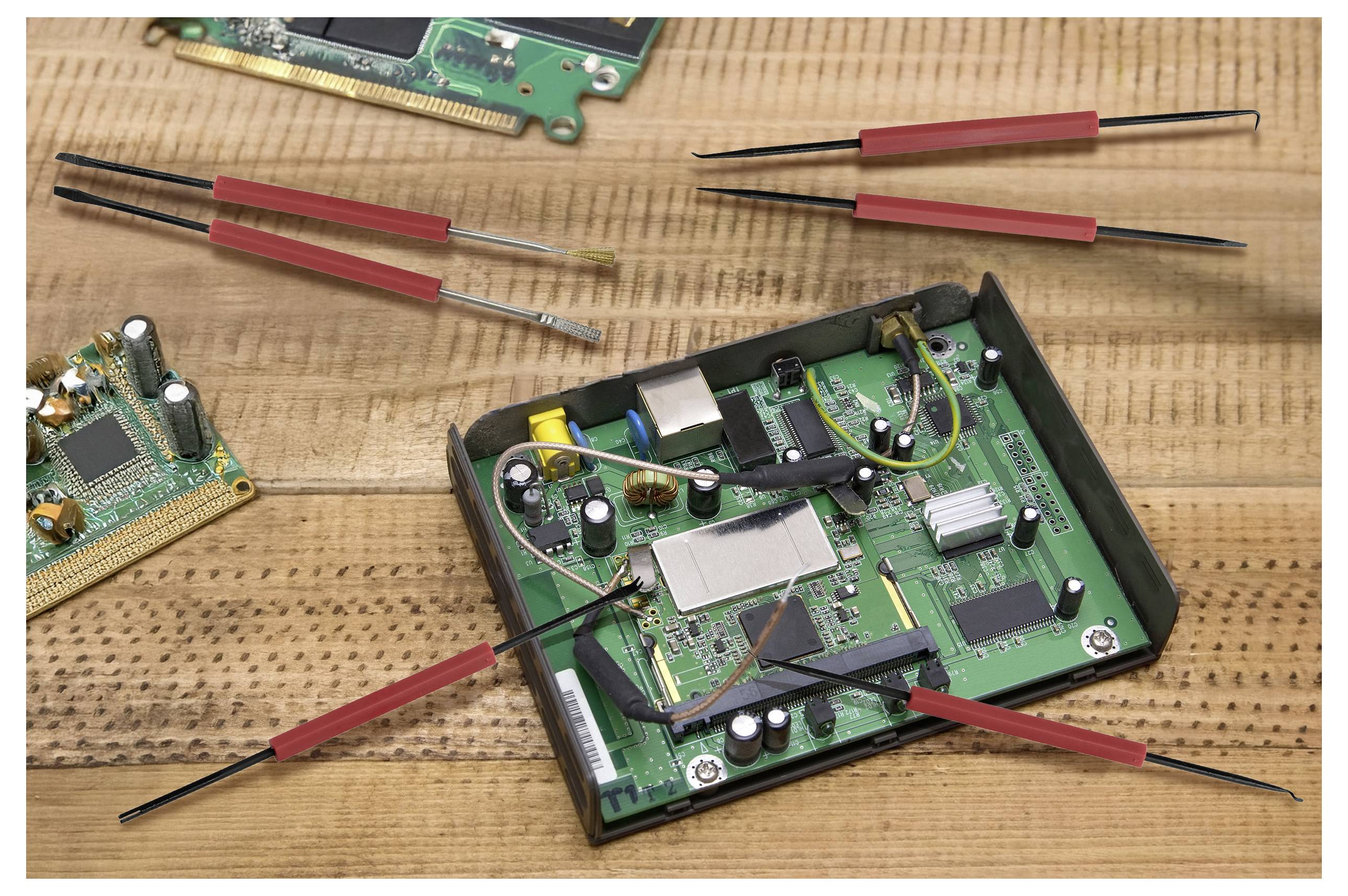 Ett öppet kretskort på ett bord med elektroniska komponenter. Flera röda och svarta pincetter är placerade runt omkring, vilket indikerar reparationsarbete.