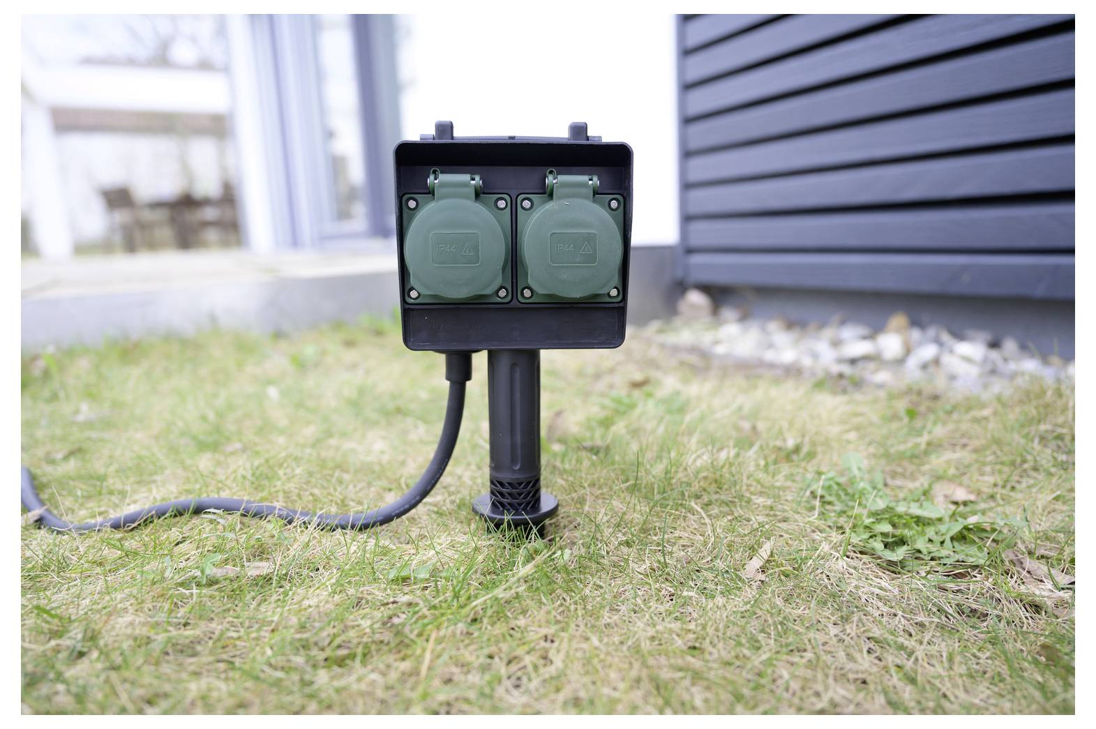 Utomhusbox för dubbel elkontakt på en kort stolpe i en gräsbevuxen trädgård, med en nätsladd ansluten nära husets exteriör.